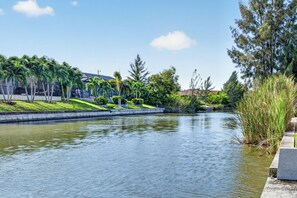A calm water body with a concrete walkway on one side, surrounded by greenery and a building in the background.
