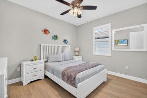 Un dormitorio con cama, mesitas de noche, ventilador de techo, decoración de peces en la pared, ventana con persianas y un cuadro colgado en la pared.