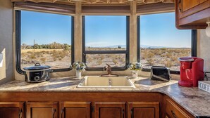 Una cocina con un lavamanos, una estufa y una cafetera. Hay dos ventanas con vistas a un paisaje desértico.