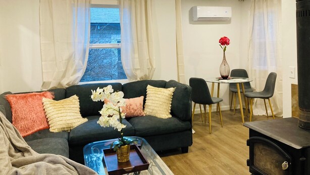 Una sala de estar acogedora con un sofá verde oscuro, una mesa de centro de vidrio y un jarrón con flores rojas. Hay un comedor con una mesa pequeña y sillas, y una estufa de leña.