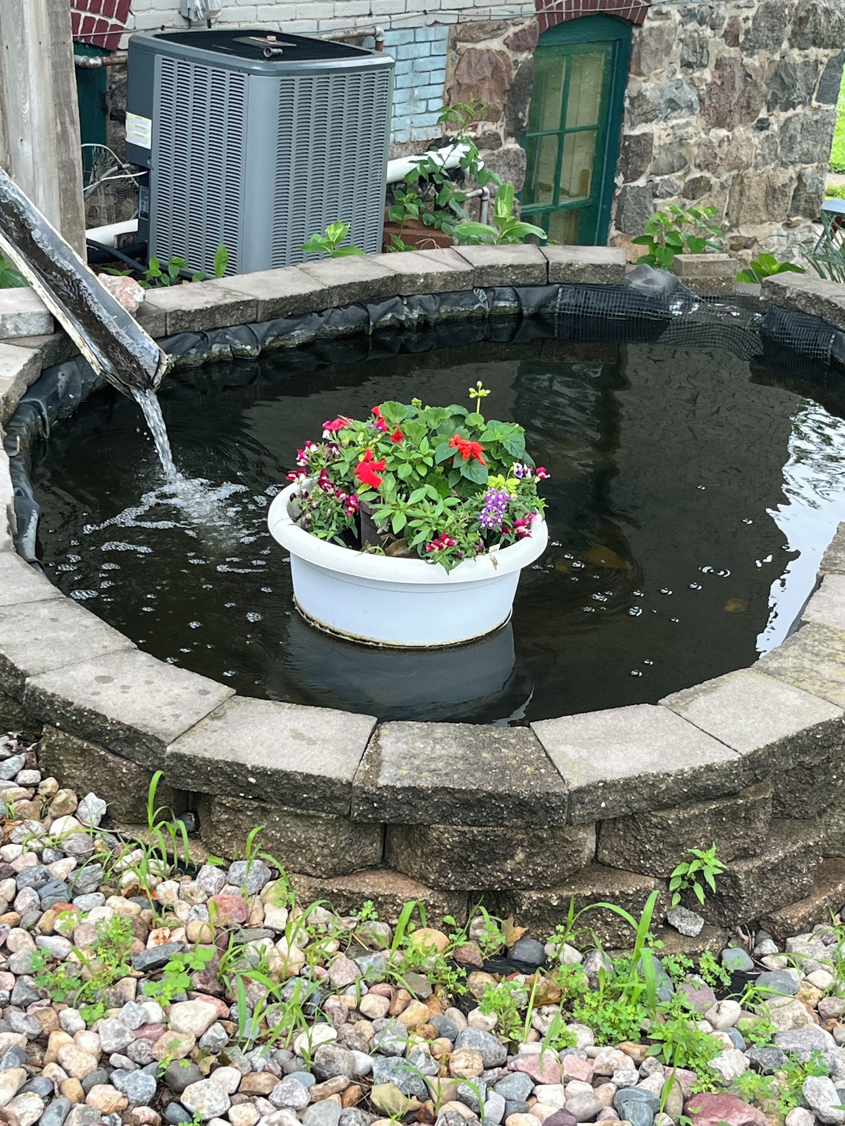 A small pond with a planter filled with flowers, surrounded by a stone edge and pebbles.