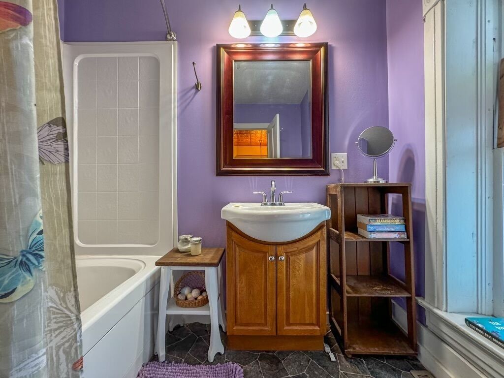 A bathroom with a white bathtub, a wooden vanity with a white sink, a mirror above the sink, and a shelf with books.