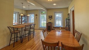 Un comedor amplio con una larga mesa de madera y seis sillas, una zona de bar con taburetes, y ventanas grandes que ofrecen luz natural.