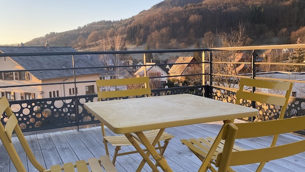 A wooden deck with yellow chairs and a table, overlooking a hilly landscape with buildings in the background.
