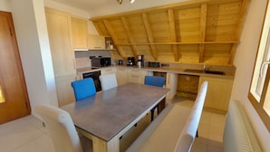 A compact kitchen with wooden beams, a dining table with chairs, a bench, and basic appliances.