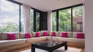 Un salon moderne avec un canapé d’angle, une table basse en verre et de grandes fenêtres donnant sur un paysage verdoyant et des bâtiments.