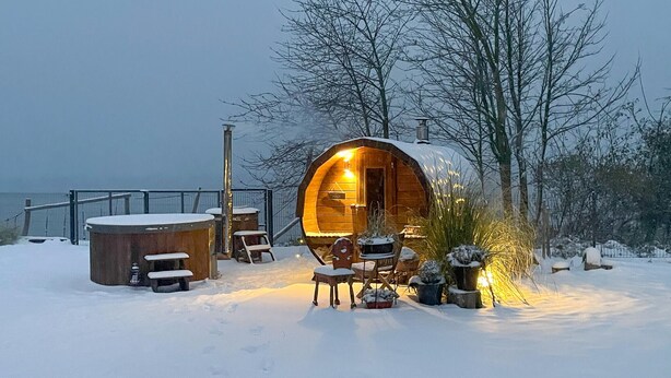A cozy wooden cabin with a warm interior light, surrounded by snow-covered ground and trees. - Beautiful home in Miedzyzdroje with sauna (Miedzyzdroje)
