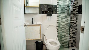 Une salle de bain avec des toilettes blanches, une poubelle noire, une cabine de douche en verre et un mur recouvert de carreaux avec un motif noir et blanc.
