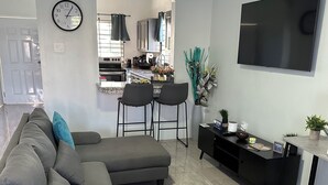A modern living room with a grey sectional sofa, two black bar stools, a flat-screen TV, and a kitchen in the background.
