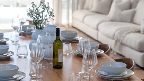 Een ronde houten eettafel gedekt met borden, kommen en wijnglazen, met een bank en gordijnen op de achtergrond.