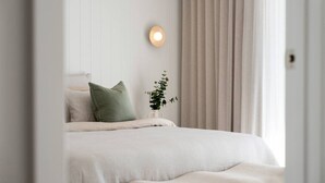 Een slaapkamer met een bed, kussens, een nachtkastje met een plant en een wandlamp.