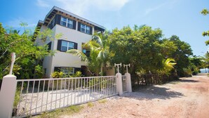 A modern two-story house with a white exterior and dark shutters, surrounded by lush greenery and a gravel driveway. - Waterfront tranquility: 3 Iguanas Villa- 1st floor Hibiscus Suite w rooftop pool (Placencia)