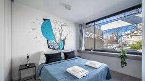 A modern bedroom with a large bed, a small bedside table, a wall mural, and a view of the city through a large window. - FLIPHAUS Malabia 1900 (Buenos Aires)