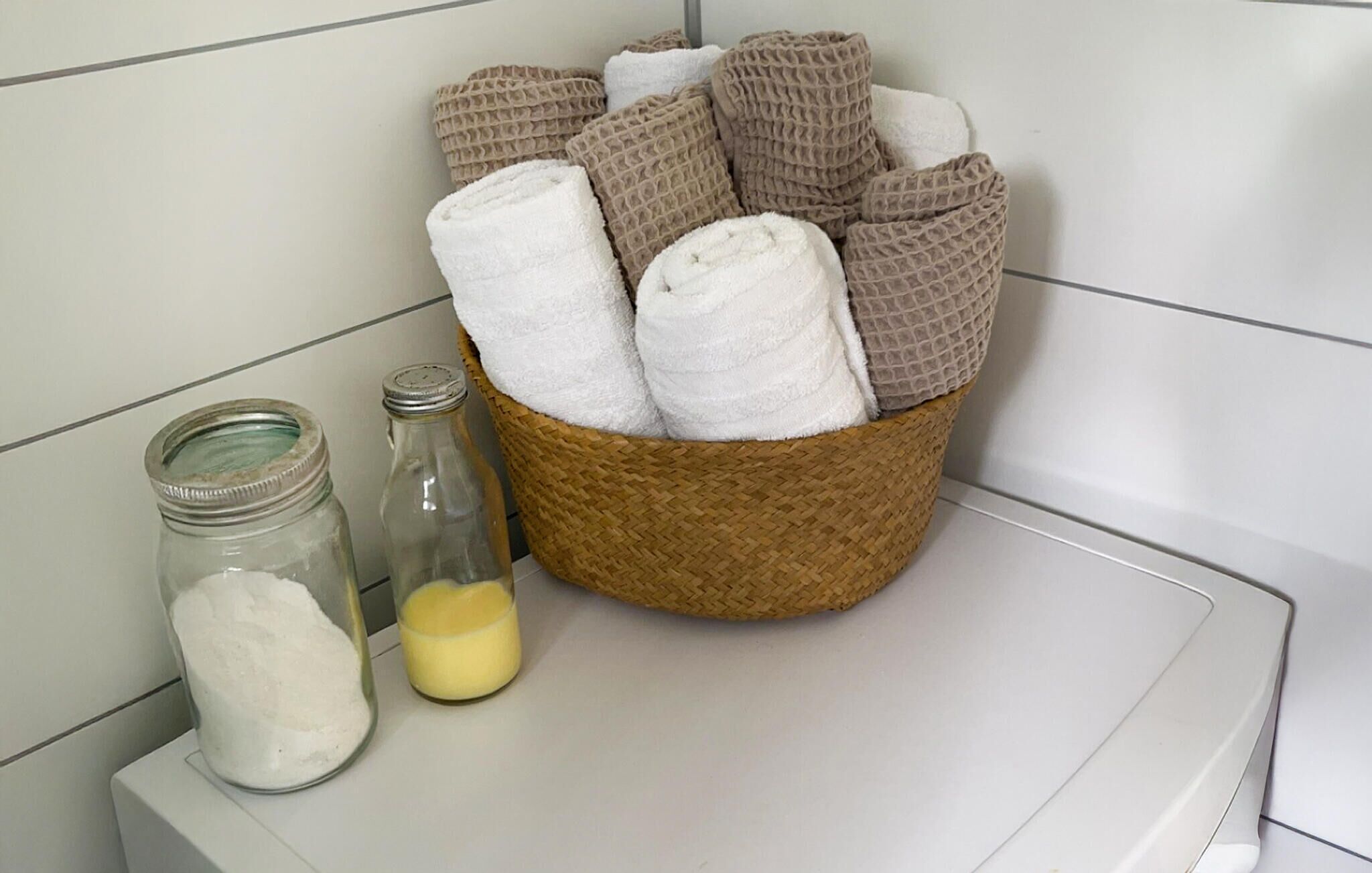 Un bagno con un cesto in vimini contenente asciugamani arrotolati, un barattolo di vetro con polvere bianca e una bottiglia di vetro con liquido giallo.
