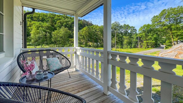 A covered porch with two wicker chairs and a small table, overlooking a road and green landscape. - 3 bedroom cozy home in Farsund (Farsund)