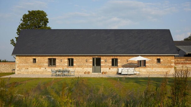 A two-story brick building with a dark roof, a front door, and a small patio area with outdoor furniture. - Gîtes de France® - La Maison d'Irène (DOUDEVILLE)