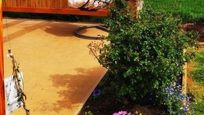 Una pérgola de madera con una hamaca colgante, un jardín con flores y un campo cercado al fondo.