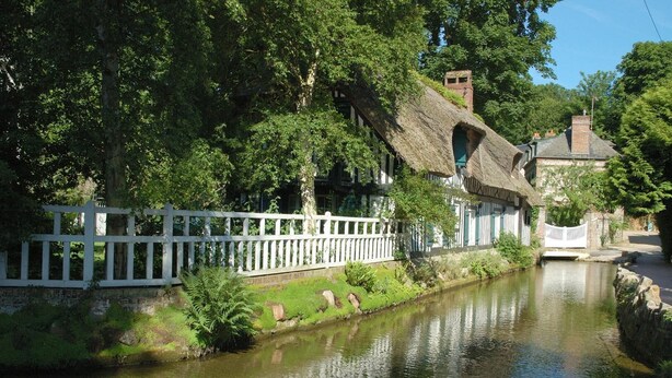 A riverside view with traditional thatched-roof cottages, a white fence, and a calm waterway. - Gîtes de France® - Le Gîte du Moulin (SAINT PIERRE LE VIEUX)