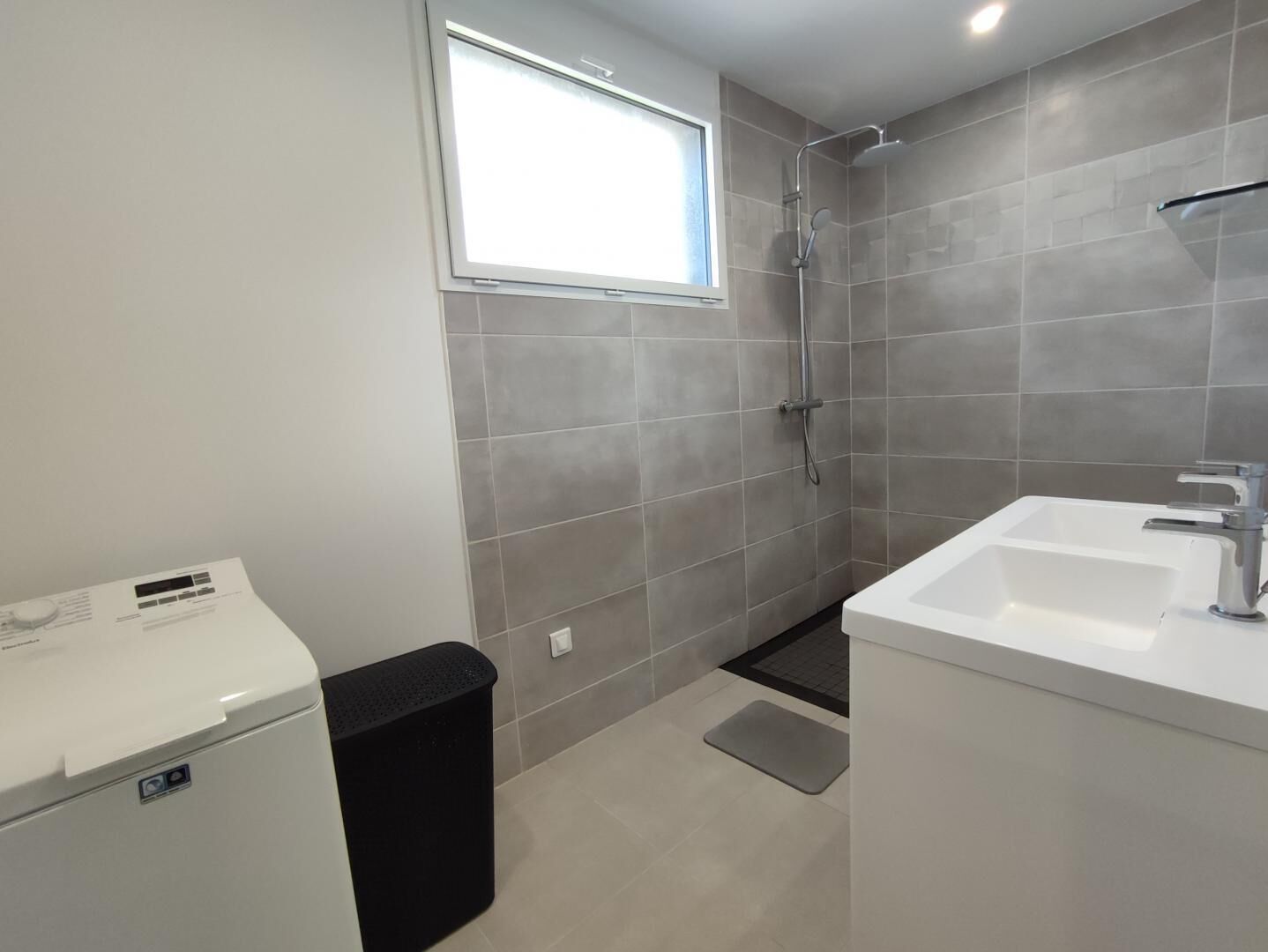 A modern bathroom with grey tiles, a window, a shower, a sink, and a trash bin.