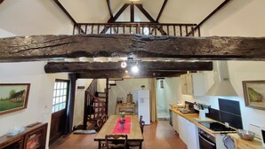 A rustic interior with exposed wooden beams, a dining area with a wooden table and chairs, a kitchen with modern appliances, and a staircase leading to an upper level.