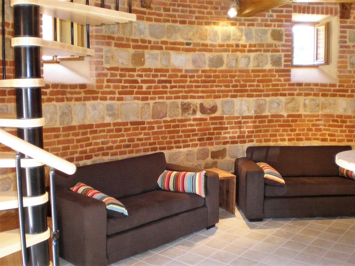 A room with a brick wall, two brown sofas with striped cushions, a spiral staircase, and a small window.