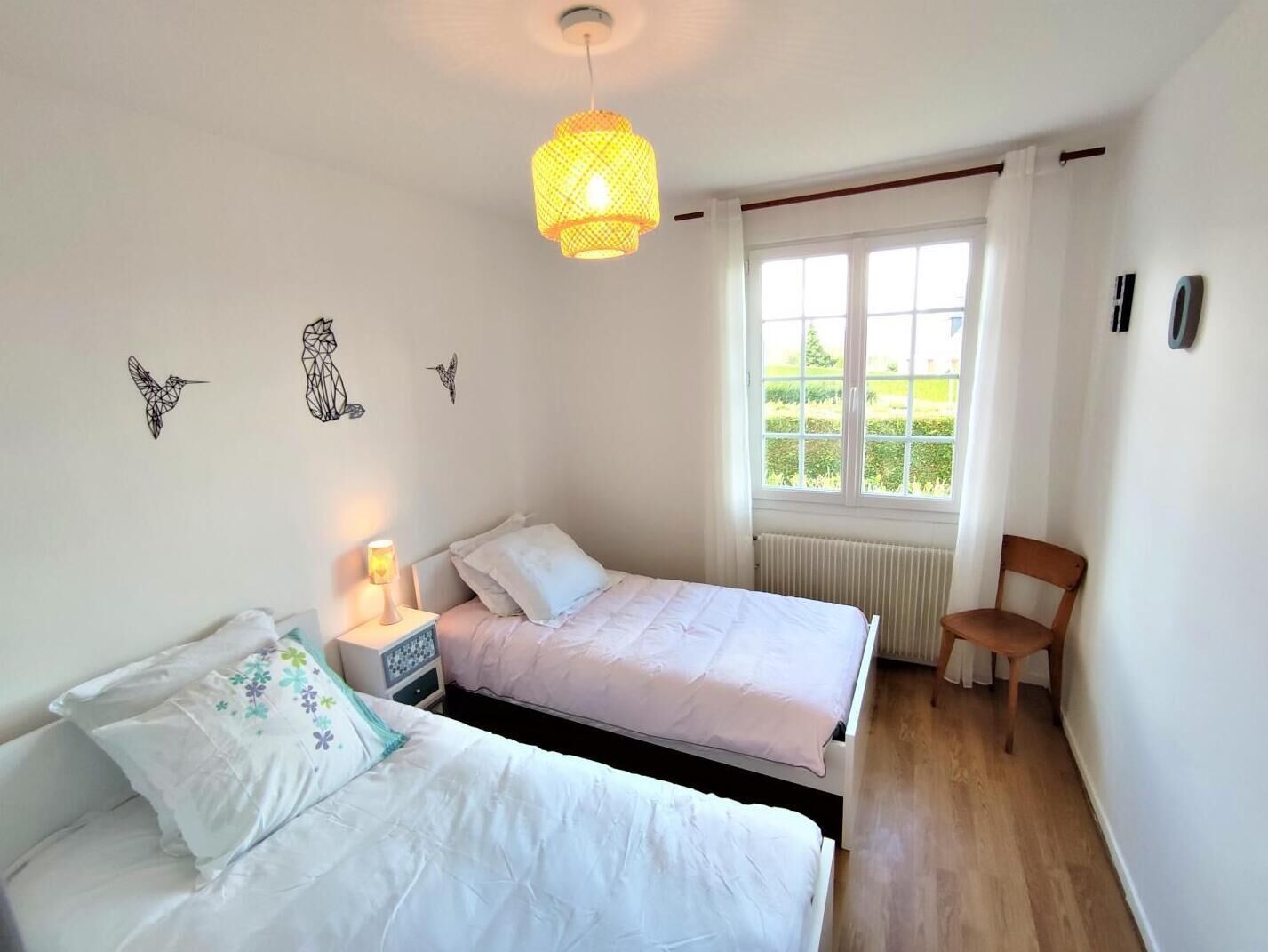 A bedroom with two beds, a window with a view of greenery, a hanging light fixture, and wall art of birds.