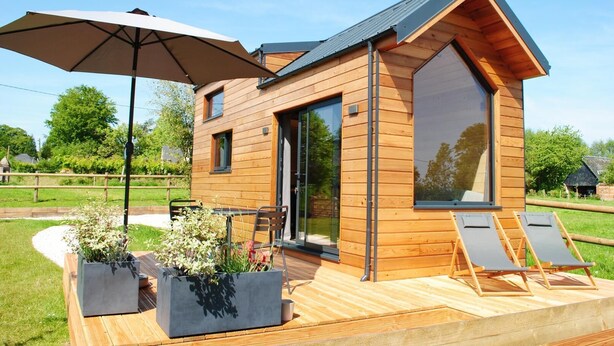 Eine Holzhütte mit einer großen Schiebeglastür, einer Terrasse mit Liegestühlen und einem Sonnenschirm.