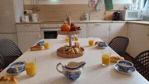 Ein Frühstückstisch mit einer mehrstöckigen Obstschale, Croissants und Orangensaft auf einer weißen Tischplatte in einer modernen Küche mit weißen Schränken und einer schwarzen Dunstabzugshaube.