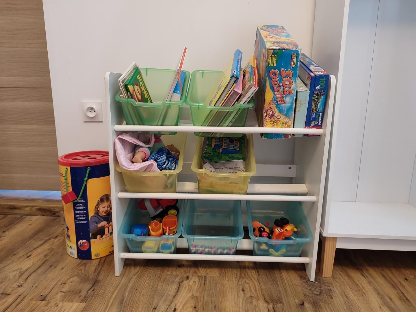 Une étagère blanche, dotée de plusieurs bacs et compartiments de rangement, dont certains contiennent des livres et des jouets.