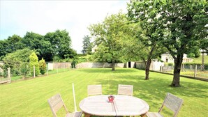 A round wooden table with six chairs in a backyard with a well-maintained lawn and trees. - Gîtes de France® - Between Sea and Earth (BAILLY EN RIVIERE)