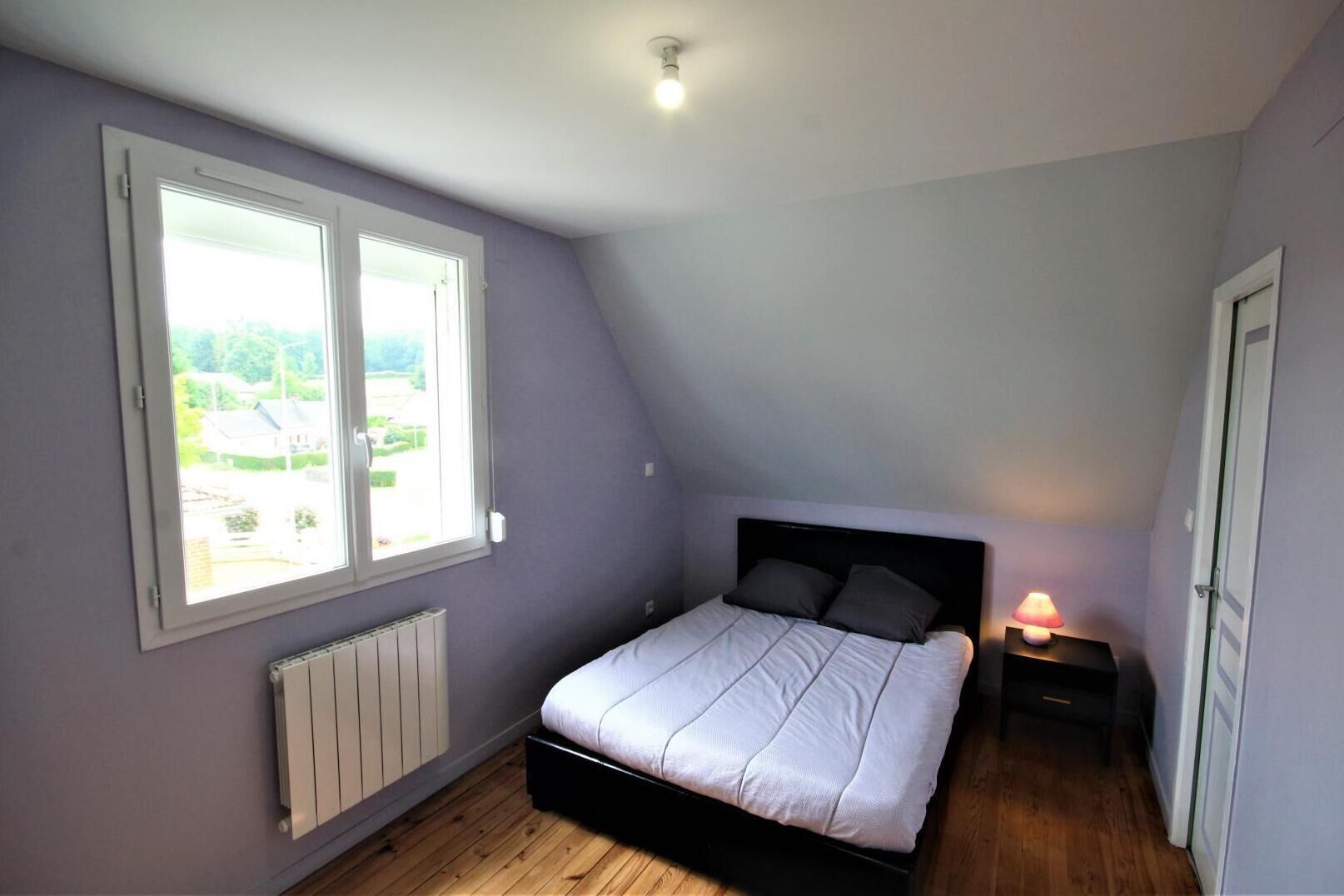 Een slaapkamer met een schuine kap, een bed met hoofdeinde, een nachtkastje met lamp en een raam met uitzicht op groen.
