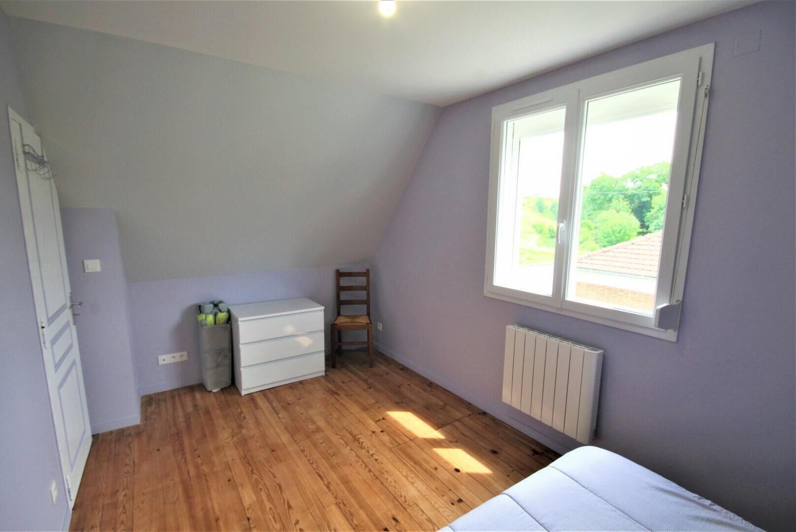 Una habitación con techo inclinado, ventana con vistas a la vegetación, un radiador y suelo de madera.