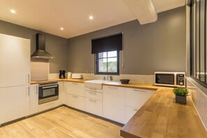 A modern kitchen with white cabinets, a built-in oven, a microwave, and a window with a black roller blind. - Gîtes de France® - Gîte du domaine de Grosmesnil (COTTEVRARD)