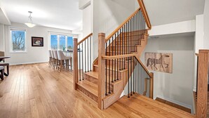 A spacious living area with a wooden staircase, a dining area with chairs, and a kitchen visible through an open door.