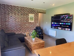 A living room with a brick wall, a black leather sofa, a wooden coffee table with a plant, a black leather chair, and a mounted TV displaying streaming services.