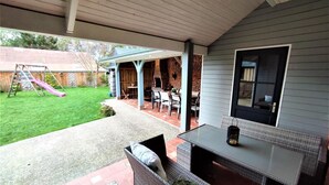 Un patio couvert avec un sol en briques, des meubles d’extérieur, un barbecue et, au fond, un ensemble de jeux pour enfants.