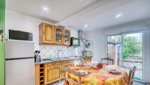 A kitchen with wooden cabinets, a microwave, and a refrigerator. There is a dining table with a tablecloth and chairs. - Gîtes de France® - Chez Gigi (LES LOGES)