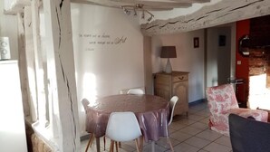 A dining area with a table covered by a plastic tablecloth, surrounded by chairs. There is a wooden cabinet with a lamp on top and a wall with a decorative quote. - Gîtes de France® - Le Petit Gîte (ERMENOUVILLE)