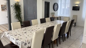 A dining area with a long table, chairs, a potted plant, a clock, and a TV on the wall. - Gîtes de France® - Gîte du Loup Vert (JUMIEGES)