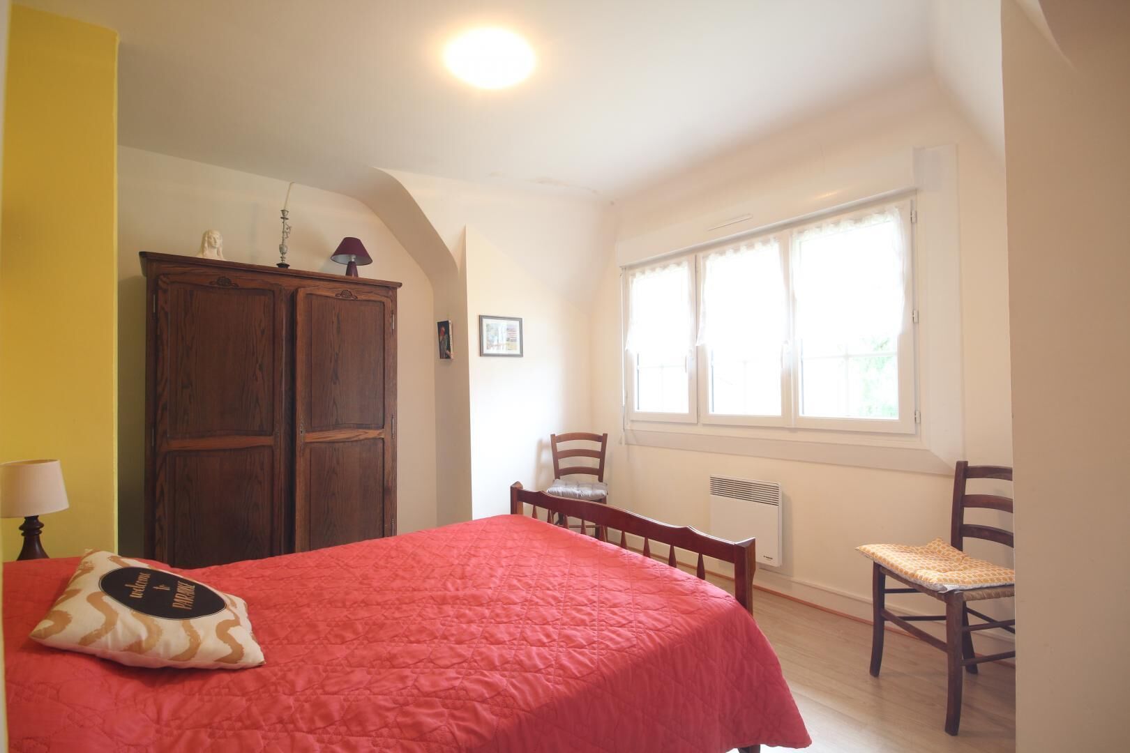 Une chambre à coucher avec un lit, une armoire en bois, une chaise, une petite table et une fenêtre avec des rideaux.