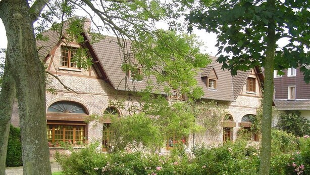 A traditional brick house with a sloped roof, large windows, and a well-maintained garden with flowering bushes.