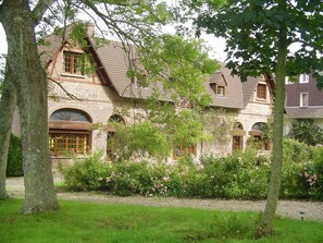 A traditional brick house with a sloped roof, large windows, and a well-maintained garden with flowering bushes. - Gîtes de France® - Les Rosiers (EU)