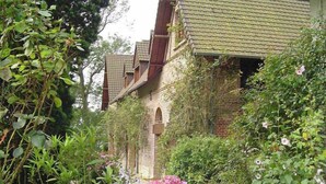 A brick building with a tiled roof, surrounded by lush greenery and flowering plants.