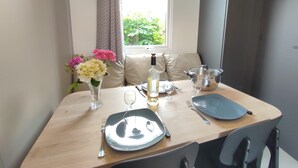 Una mesa de comedor dispuesta con platos, cubiertos y una botella, con una ventana y cortinas al fondo.