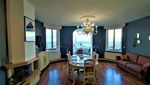 A spacious living room with a dining area, large windows offering a view of the sea, a chandelier, and a cozy sofa. - Gîtes de France® - Le Côtier (FECAMP)