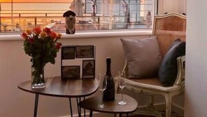 A room with a view of a sunset over a body of water, a small table with a vase of flowers, a bottle of wine, and a wicker chair with cushions. - Gîtes de France® - Le Côtier (FECAMP)