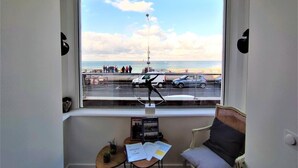 A room with a large window overlooking a beach, a chair, a small table with a notebook and papers, and a wall-mounted shelf with books. - Gîtes de France® - Le Côtier (FECAMP)
