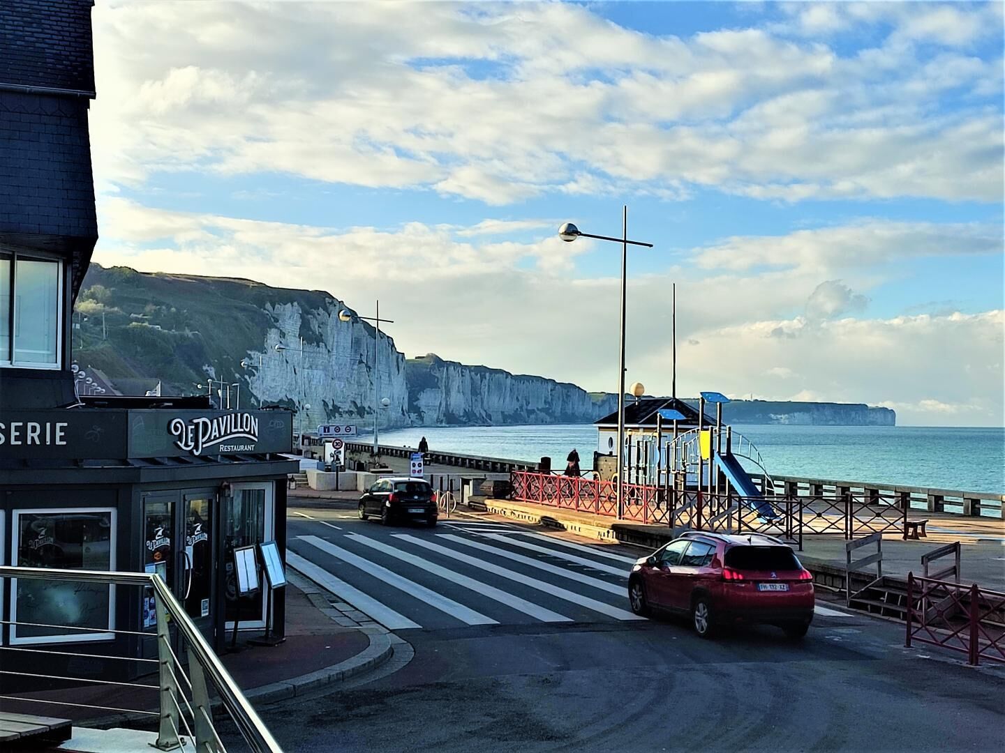 A coastal road with a restaurant named "Le Pavillon" on the left, a pedestrian crossing, and a cliffside view of the sea.