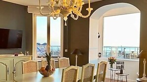 A dining area with a chandelier, round table, and chairs. There is a television on the left and a balcony with a view. - Gîtes de France® - Le Côtier (FECAMP)