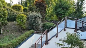A wooden deck leading to a staircase with a glass balustrade, surrounded by a well-maintained garden with various plants and a palm tree. - Gîtes de France® - At the crossroads (BERNEVAL LE GRAND)